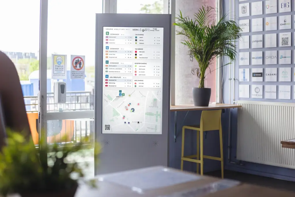 Der VEOMO-Mobilitätsmonitor im Einsatz – Co-Working Space. Der Bildschirm zeigt Abfahrtszeiten von ÖPNV, Sharing-Angebote und eine Umgebungskarte.